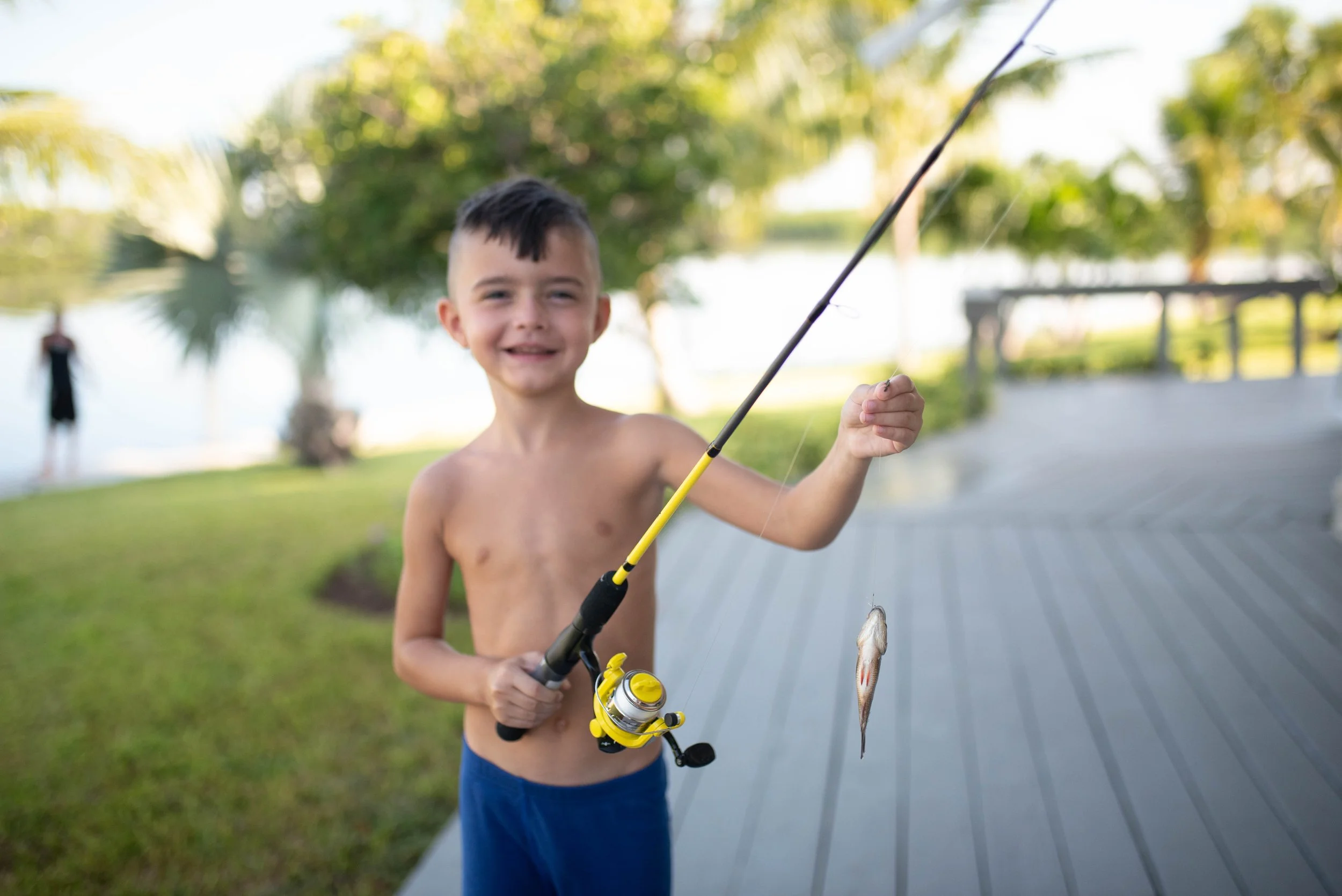 A young boy with short dark hair, shirtless, holding a fishing rod with a small fish hanging from the line, smiling on a wooden dock near a lake with greenery in the background.
