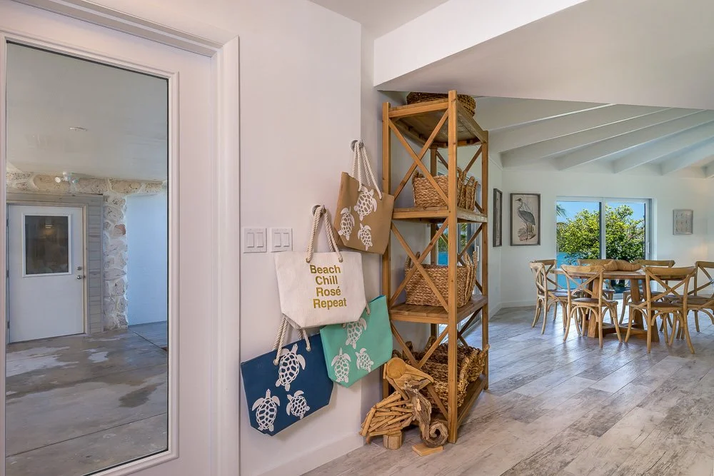 Interior view of a bright, coastal-style home with a dining area, wooden shelving with baskets, hanging tote bags with sea turtle prints and one with the text "Beach Chill Rosé Repeat," large window with greenery outside, and a sliding glass door lea
