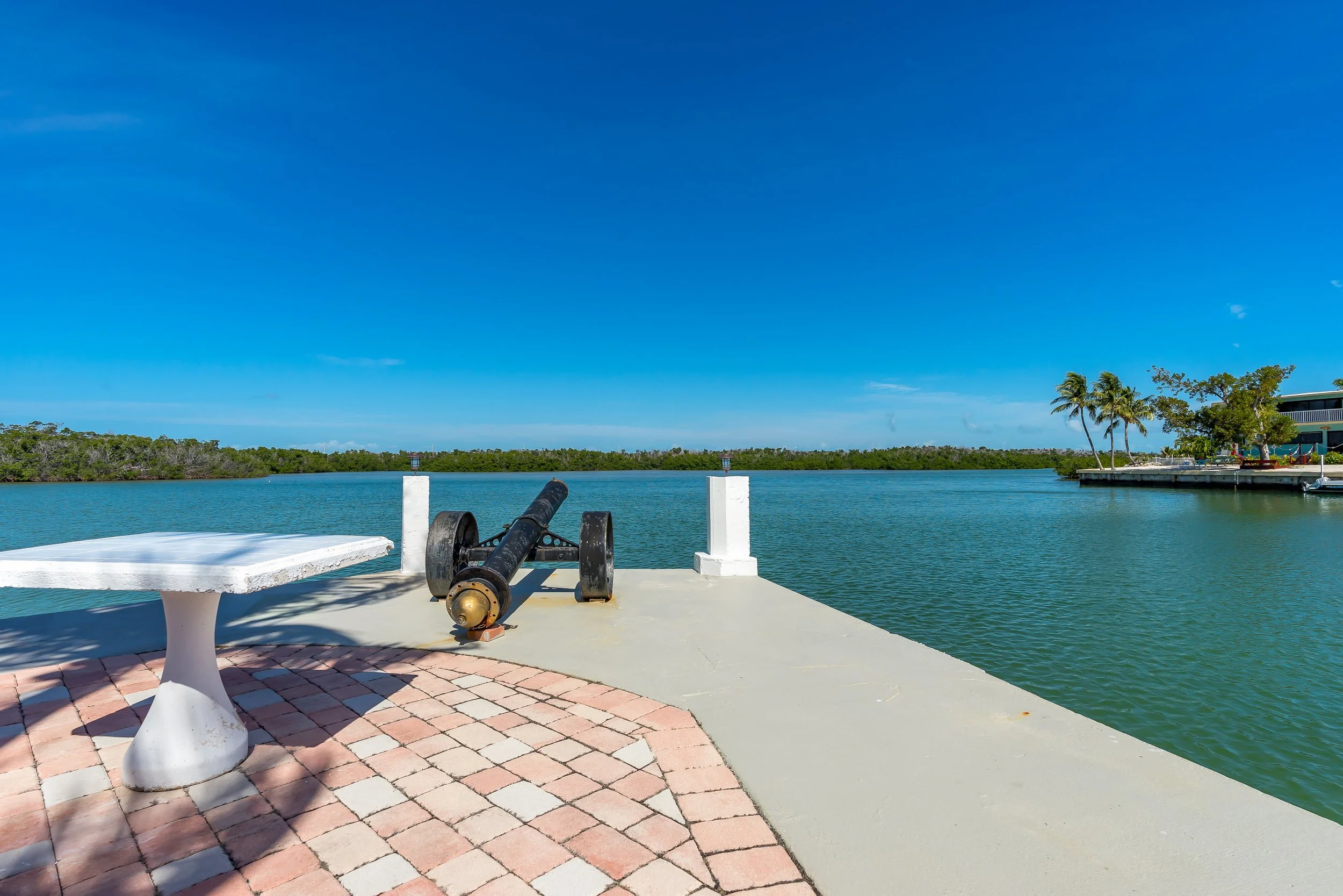 A waterfront scene with a cannon on a dock, a white statue-like bench, brick paving, water, trees, and a building with palm trees against a clear blue sky.