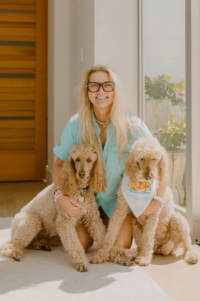 A woman with long blond hair, glasses, and a turquoise shirt kneeling indoors with two poodles, one with a uniform bandana, near a glass door with plants outside.