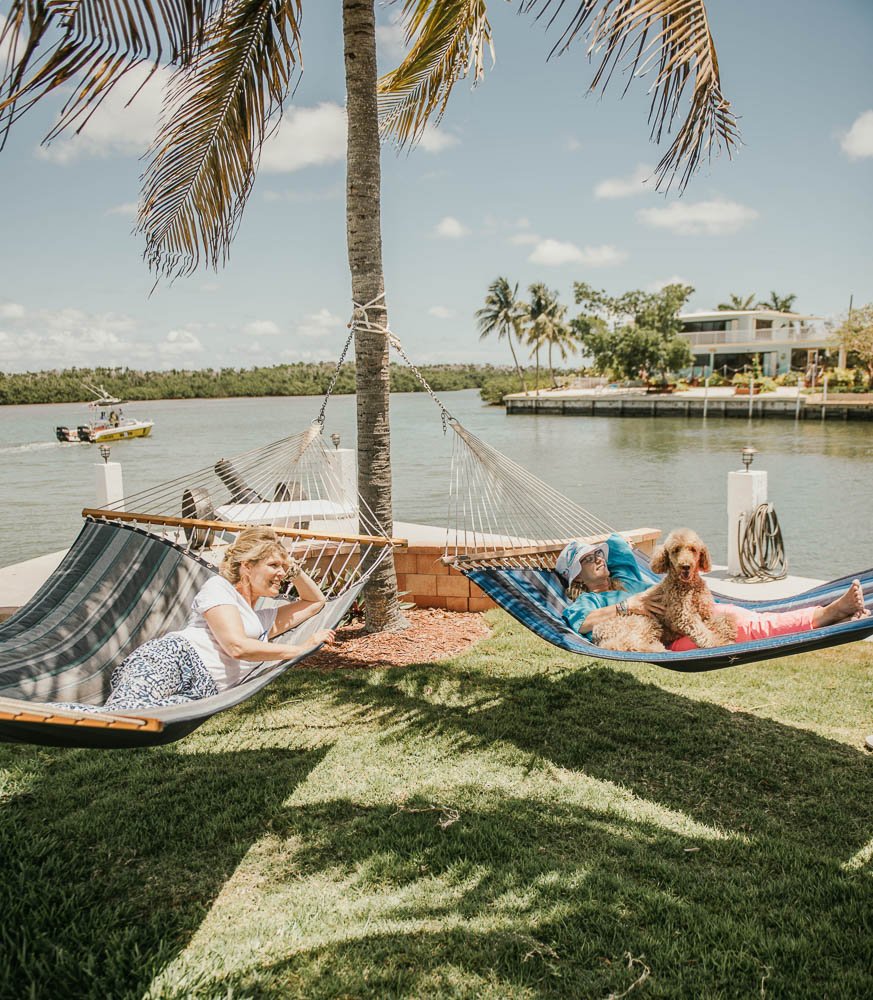Two women relaxing in hammocks tied to a palm tree by the water, with a woman and a dog in the hammock on the right.