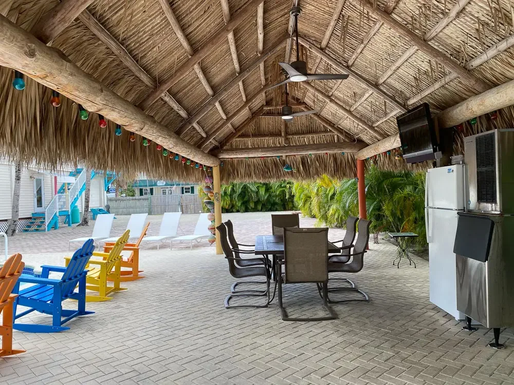 An outdoor poolside area with colorful Adirondack chairs, a thatched roof cabana with a ceiling fan, a dining table with chairs, and a volleyball net in the background, surrounded by lush green plants.