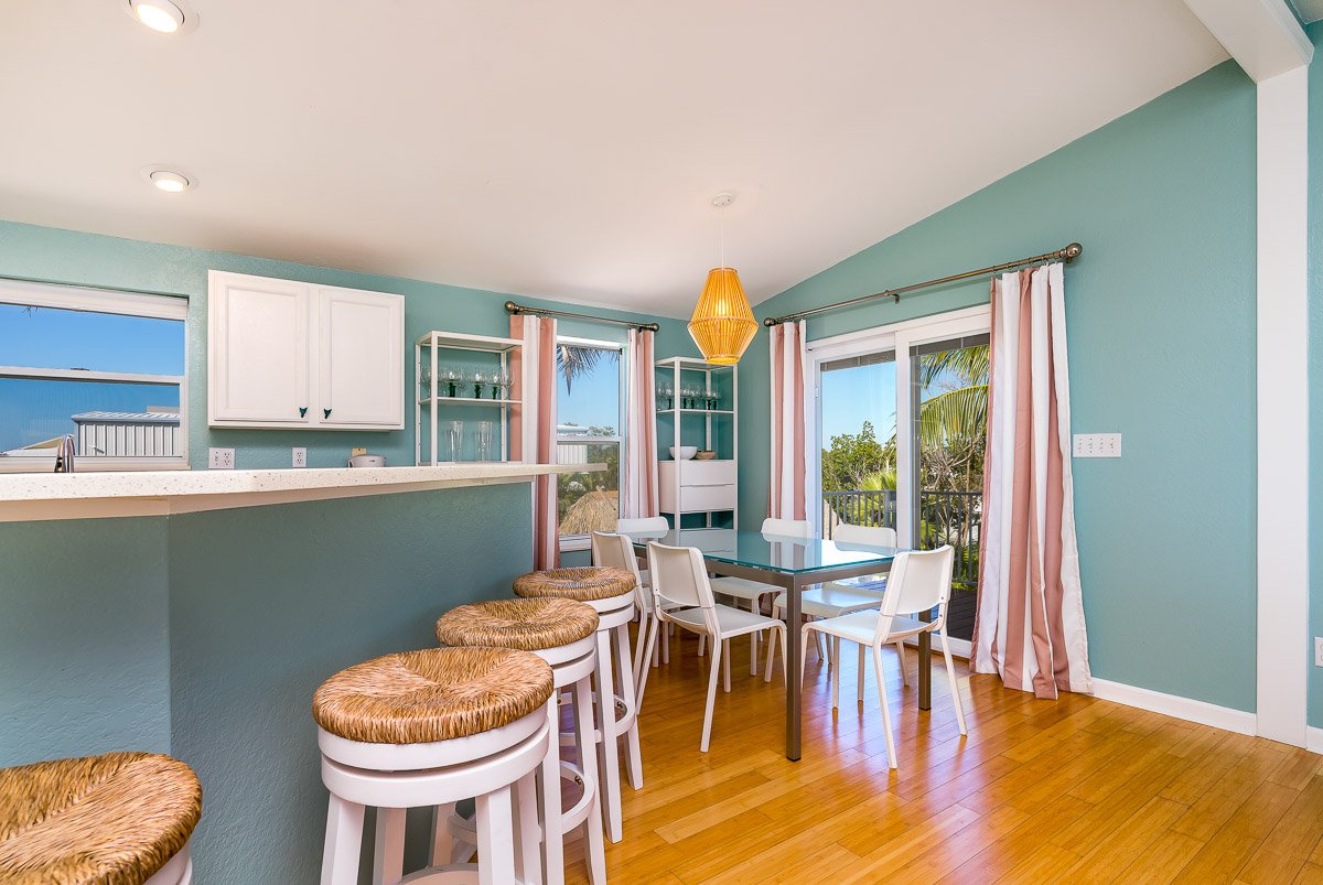 Dining area with a table and chairs, a breakfast bar with stools, and large windows with curtains overlooking a balcony and outdoor greenery.