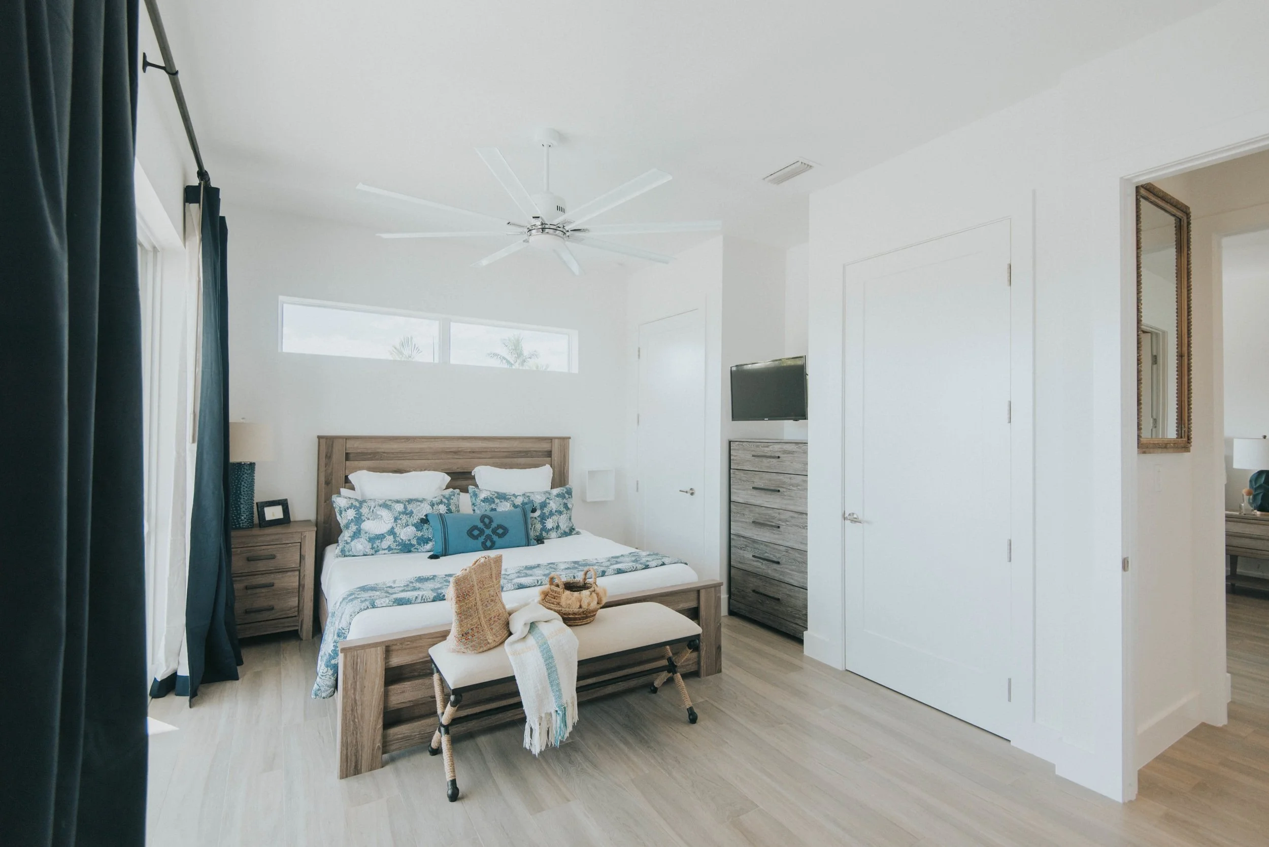 Bright bedroom with a wooden bed, blue and white bedding, dresser with a television, and a bench at the foot of the bed with baskets and textiles.