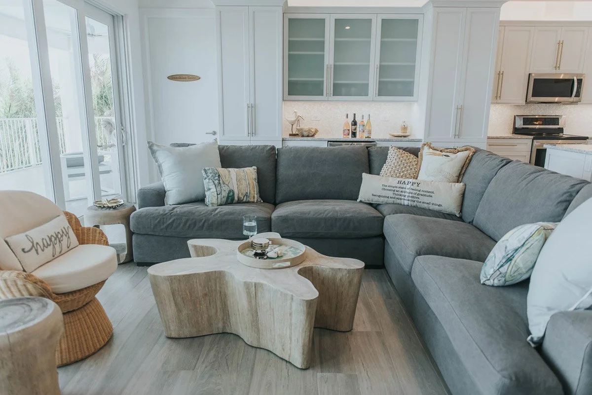 Living room with a large L-shaped gray sectional sofa, a white and beige coffee table, pillows with decorative text and patterns, and an armchair with a pillow that says "Happy". In the background, there is a white kitchen with glass cabinet doors an