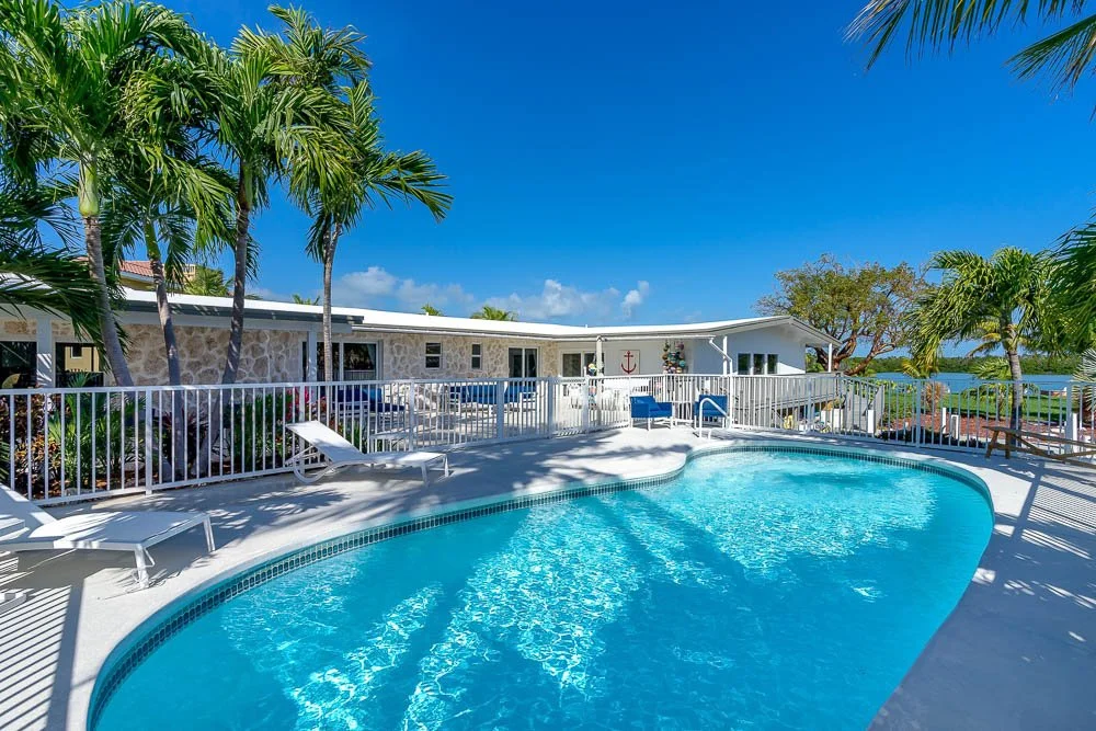 A backyard with a swimming pool, surrounded by palm trees, lounge chairs, and a white fence, with a house in the background and a clear blue sky.