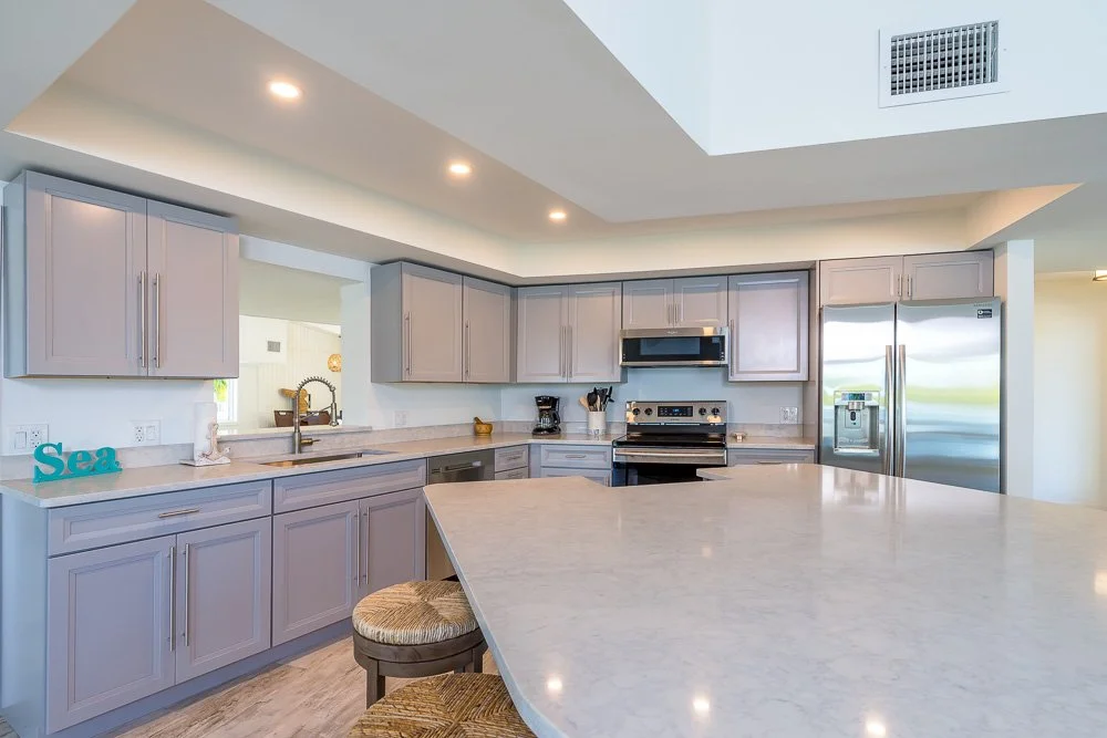 Modern kitchen with gray cabinets, stainless steel appliances, and a large island with a marble countertop. Recessed ceiling lights and a view of the outdoors through a window.