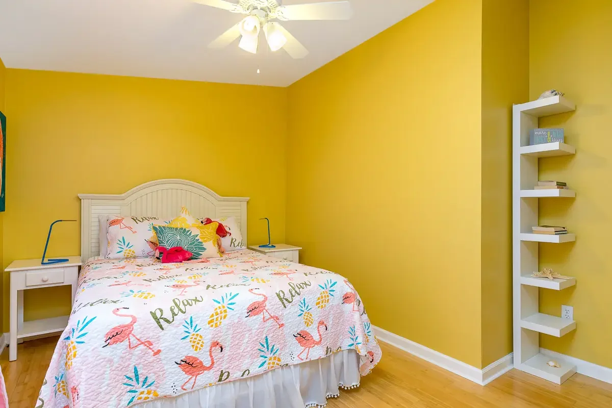 A bedroom with yellow walls, a white bed with flamingo and pineapple patterned bedding, a white headboard, two white nightstands, a ceiling fan with lights, and a white corner shelf with books and decorations.