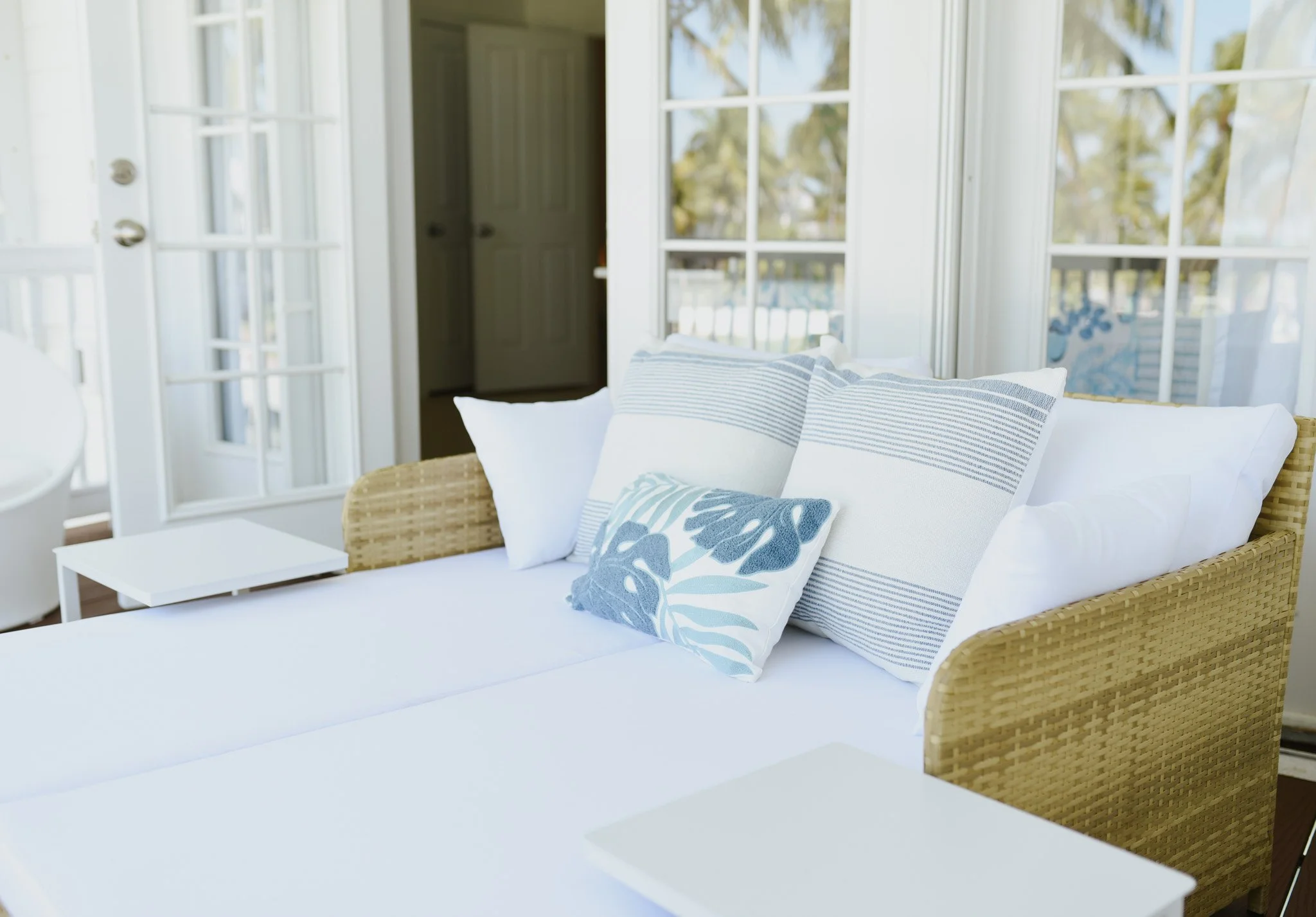 Indoor scene of a wicker sofa with white cushions and blue striped and floral pillows, near large windows with sunlight.