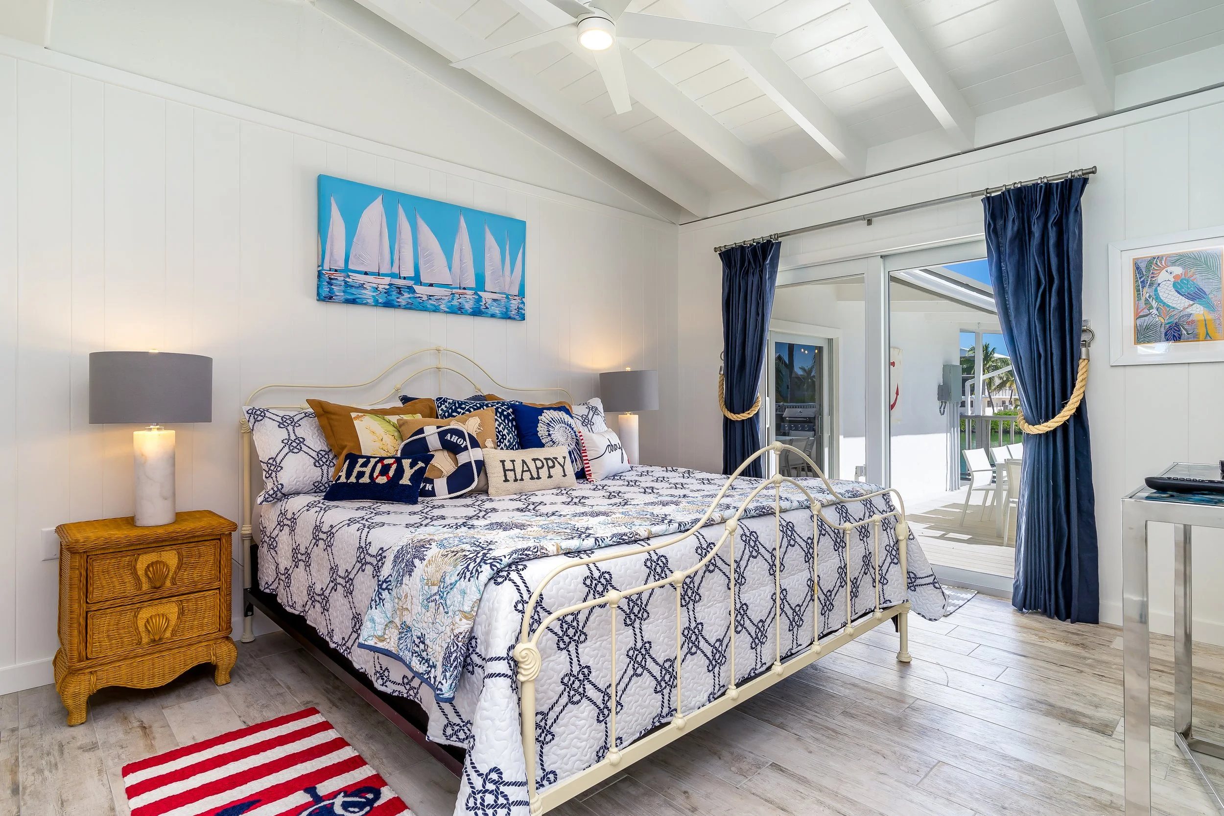 A bedroom with a white iron bed, nautical-themed pillows, a wicker nightstand, and a sliding glass door with blue curtains leading to an outdoor patio.
