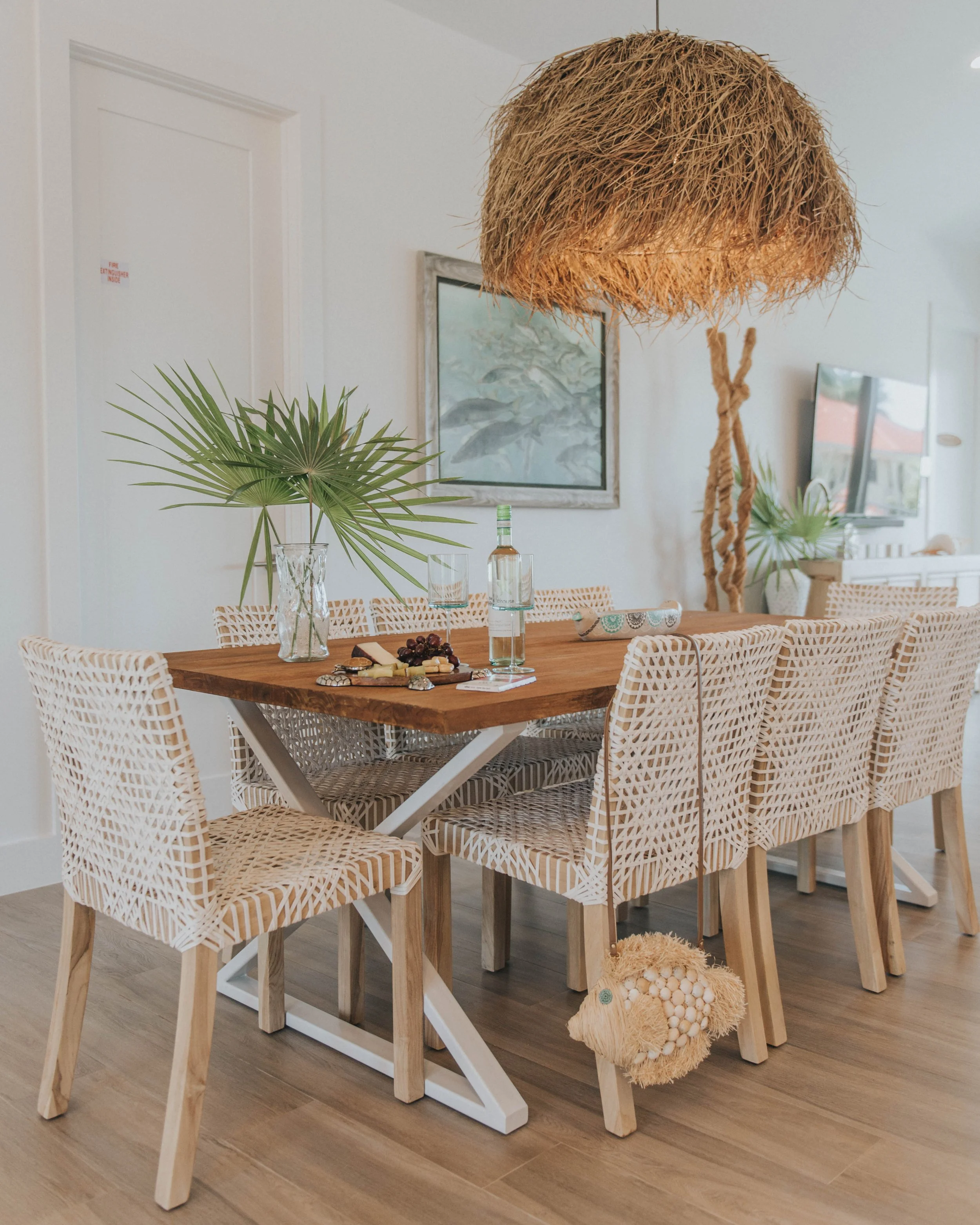 Dining room with a wooden table, surrounded by white woven chairs, a large leaf in a glass vase, wine bottle, glasses, and snacks on the table. A large woven pendant light hangs above, and there is a decorative dog-shaped bag hanging from one chair. 