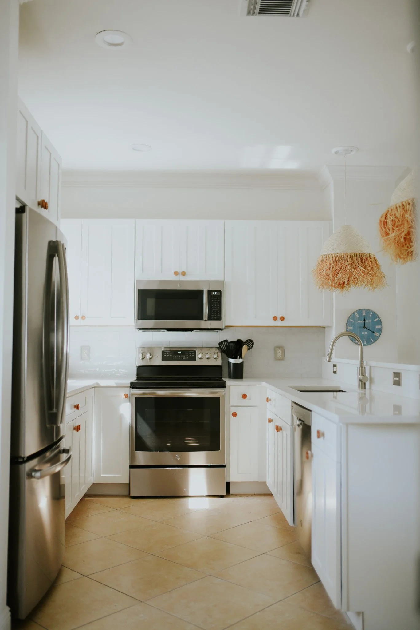 Modern kitchen with white cabinets, stainless steel refrigerator, oven, microwave, black utensil holder, blue wall clock, and two hanging pendant lights with beige fringes.