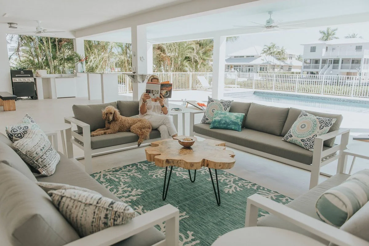 Bright, open living room with white furniture, patterned pillows, a green area rug, and a wood coffee table. A woman is sitting on a sofa reading a magazine, with a dog lying beside her. Large windows and sliding doors reveal a pool and outdoor seati