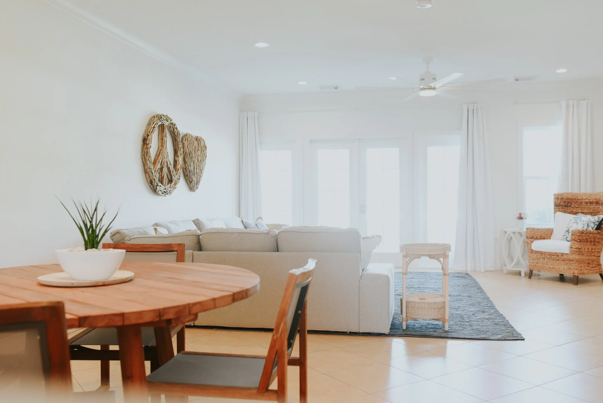 Bright living room with white walls, beige sofa, wicker armchair, small white side table, wooden dining table with chairs, and large windows with white curtains.