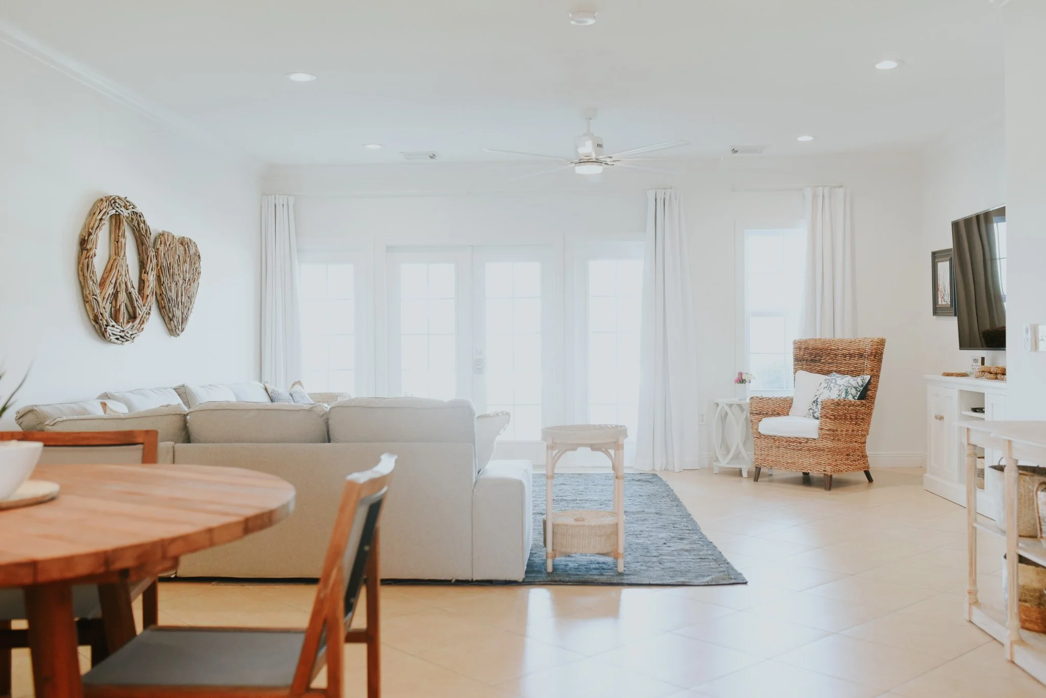 Bright living room with white walls, cream sofa, rattan armchair, glass side table, and television on white cabinet. Light-colored tiled floor and large windows with white curtains.
