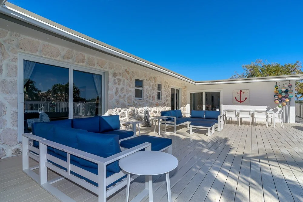 Outdoor patio with white furniture and blue cushions, stone wall house, nautical decor, clear sky, and deck chairs with colorful umbrellas.