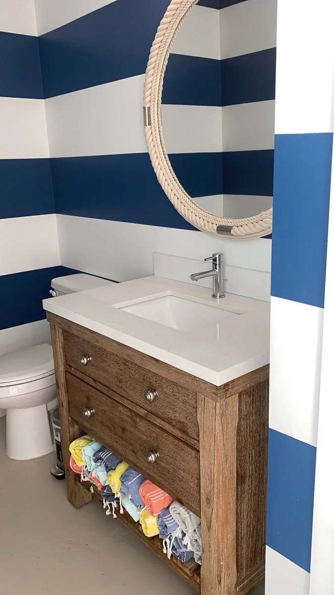 A bathroom with a striped blue and white wall, a round mirror with a woven frame, a white sink with a wooden cabinet underneath, and a toilet. The cabinet has three drawers and an open shelf filled with folded colorful towels.