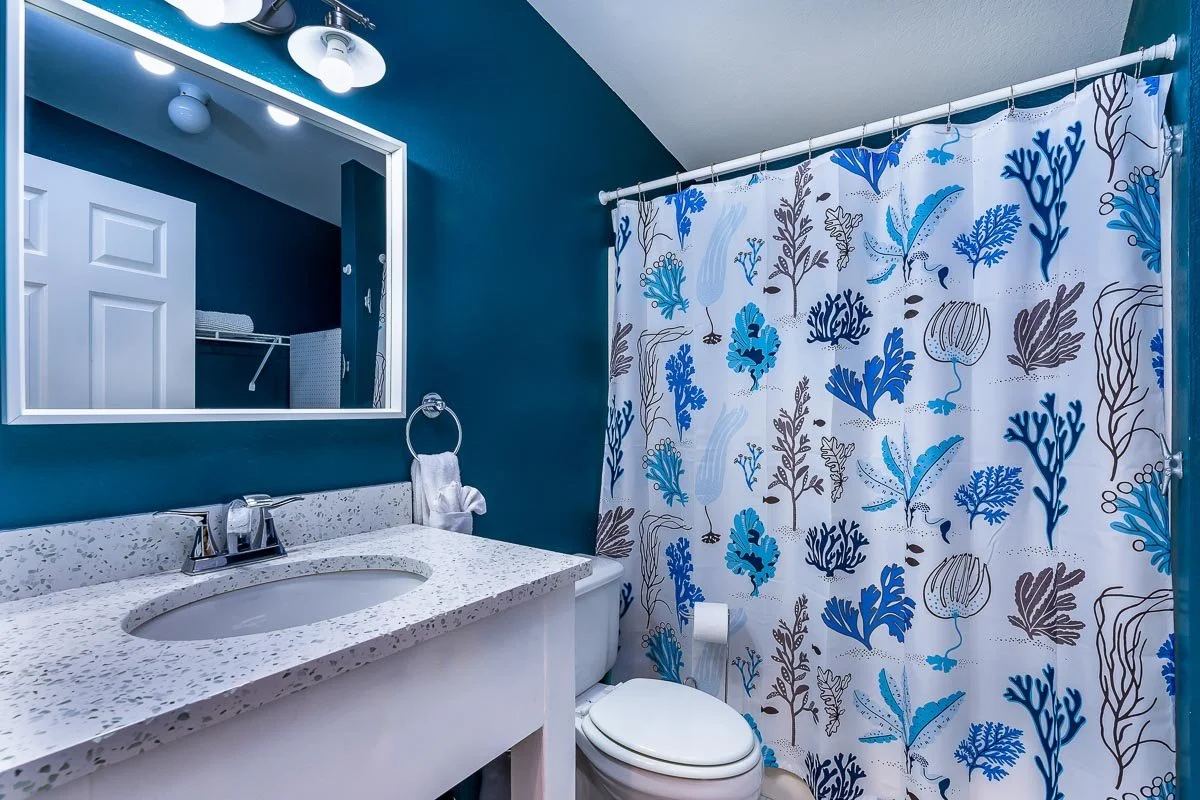 Bathroom with a white speckled countertop sink, large mirror, teal walls, shower curtain with blue, teal, and brown botanical pattern, white toilet, and towel ring with a towel.