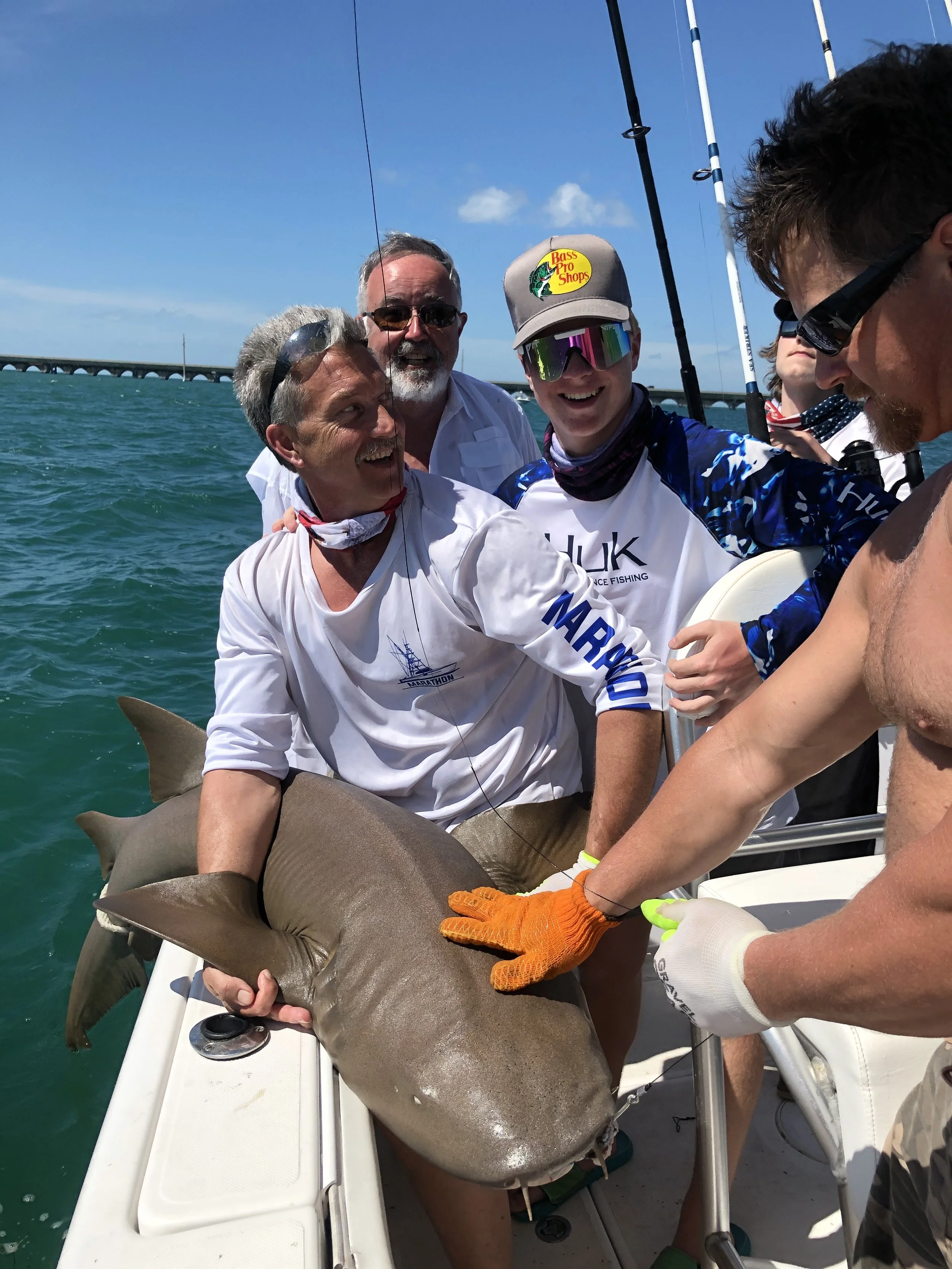 Group of smiling men on a boat holding a large fish, with a bridge in the background under a bright blue sky.