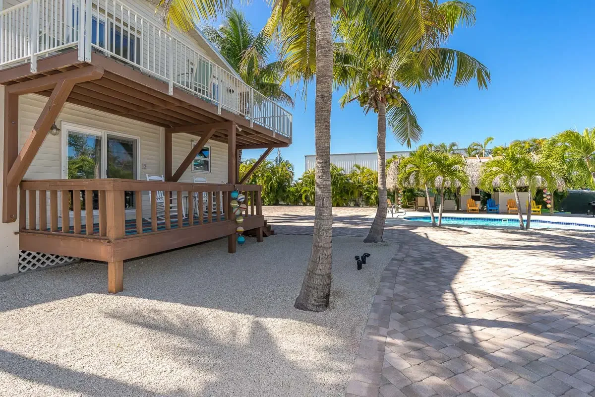 View of a backyard with a swimming pool, palm trees, a patio with brick pavers, and a two-story house with a deck.