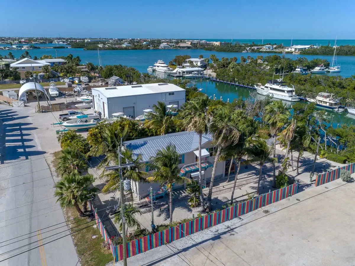 Aerial view of a marina with boats, a building, and palm trees near water, with a colorful striped fence along a street in a coastal town.