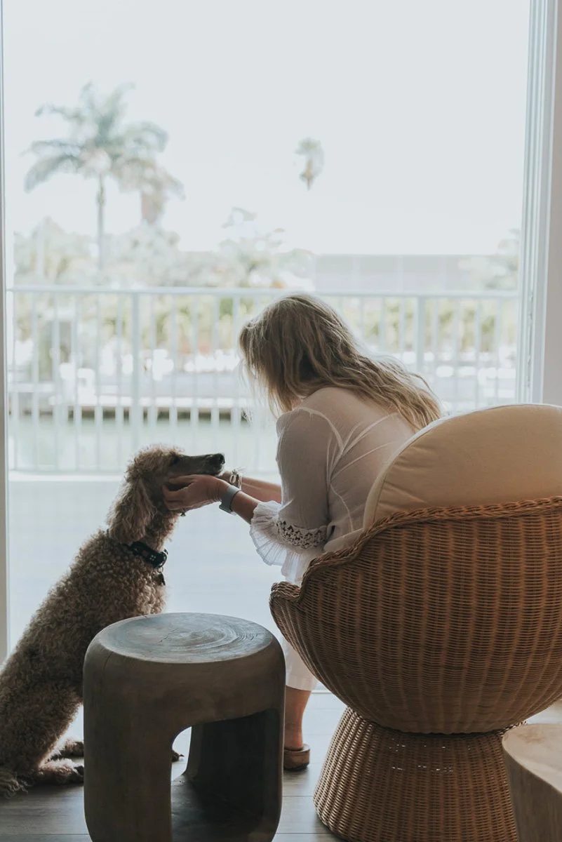 A woman sitting in a wicker chair with her dog, inside a room near a large window, petting the dog's head, with palm trees visible outside.