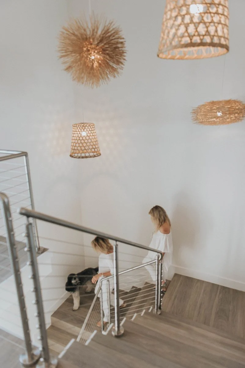 Two young girls in white dresses petting a black and white dog on a staircase with modern railing, in a well-lit interior space with decorative hanging lights and a white wall.