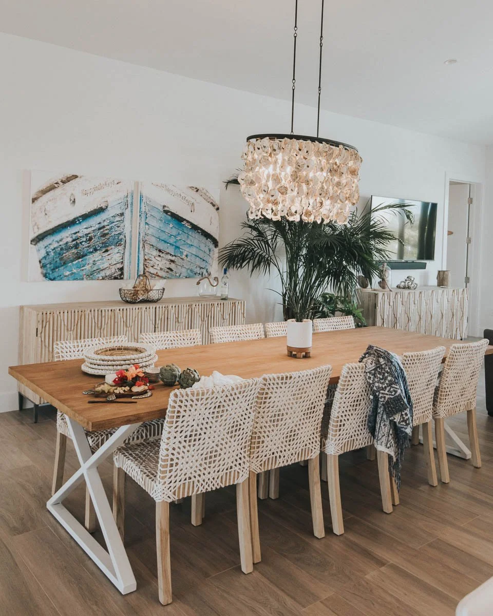 Interior of a dining room featuring a large wooden table with ten wicker chairs, a chandelier with seashell-like decor, a large leafy plant, wall-mounted TV, artwork of a boat, and a sideboard with coastal decorations.
