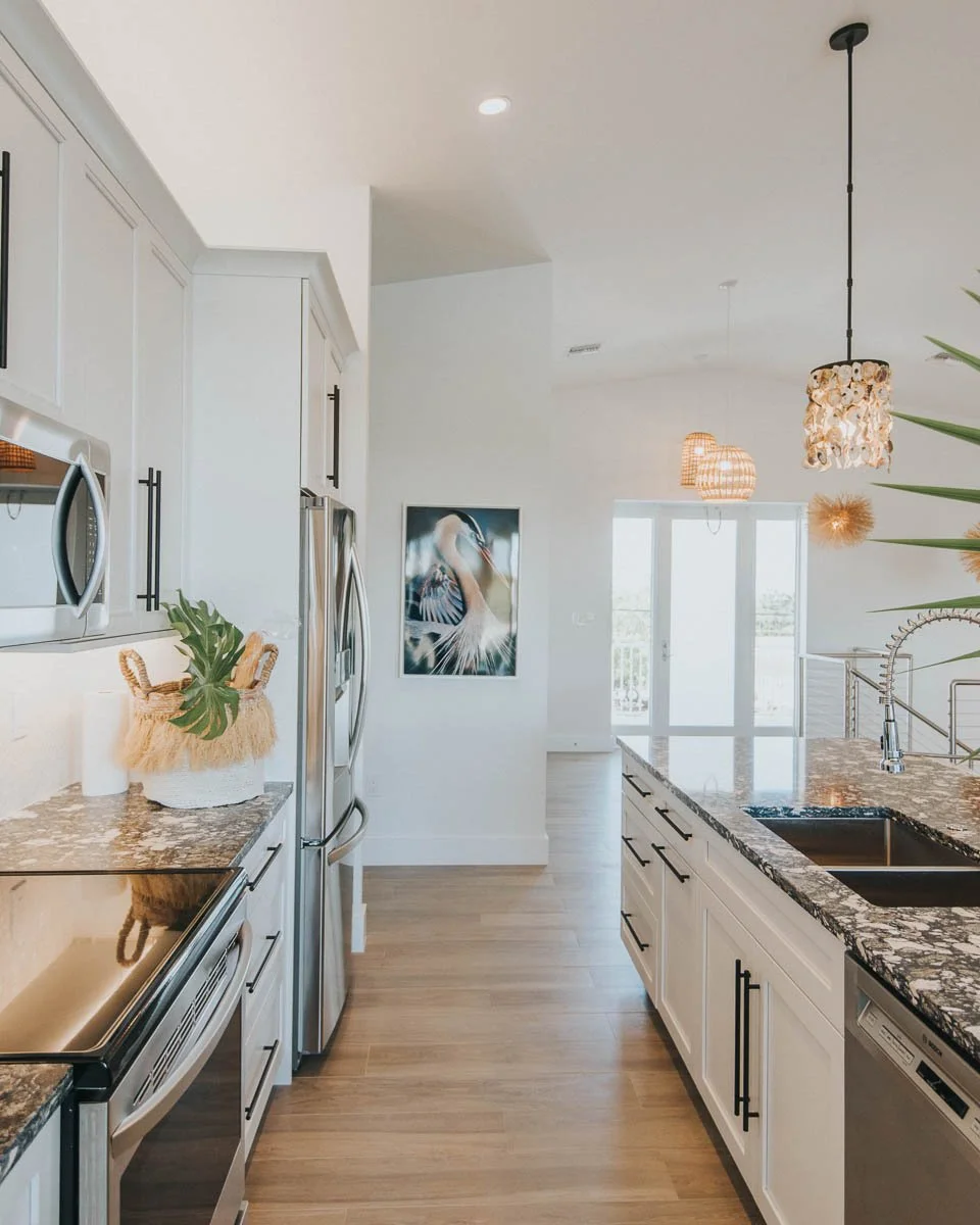 Modern kitchen with white cabinets, granite countertops, stainless steel appliances, pendant lights, and wood flooring. There is a painting of a bird on the wall and a sliding glass door in the background.