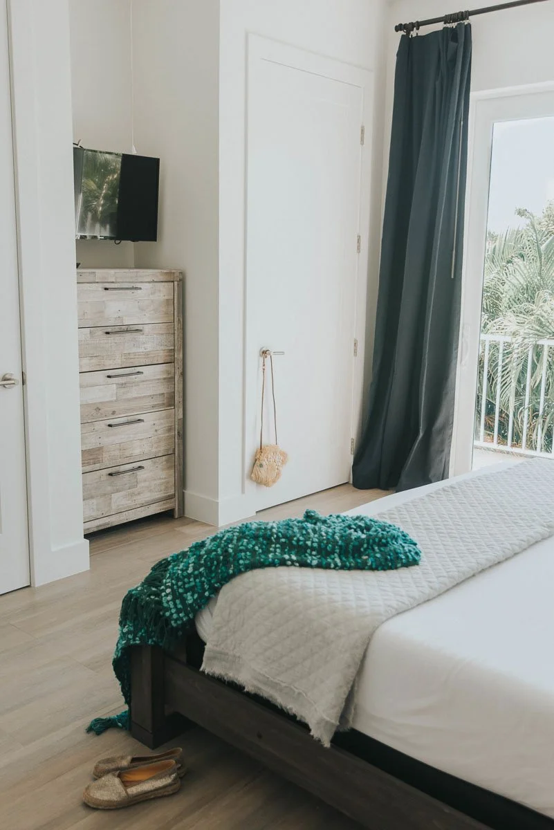 A bedroom with a white bed, textured white blanket, and teal throw at the foot. A pair of beige slippers on the light wood floor. A dark curtain on a sliding door leading to a balcony with greenery outside. A white door with a small decorative bag ha
