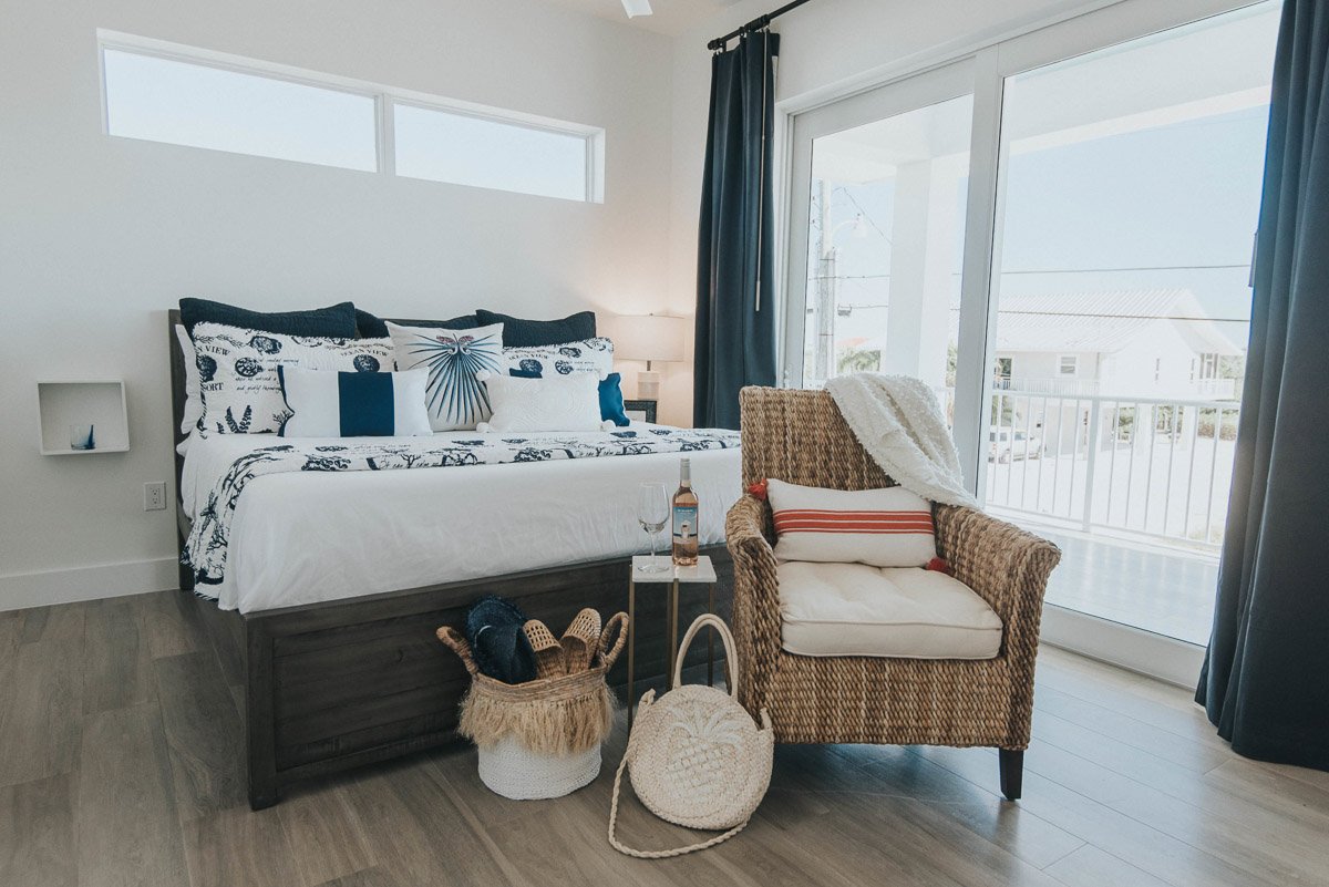 A cozy bedroom with a large bed, multiple decorative pillows, and a dark-colored headboard. There is a small side table with a wine glass and bottle. Next to the bed is a wicker armchair with a white pillow and a throw blanket. In front of the chair 