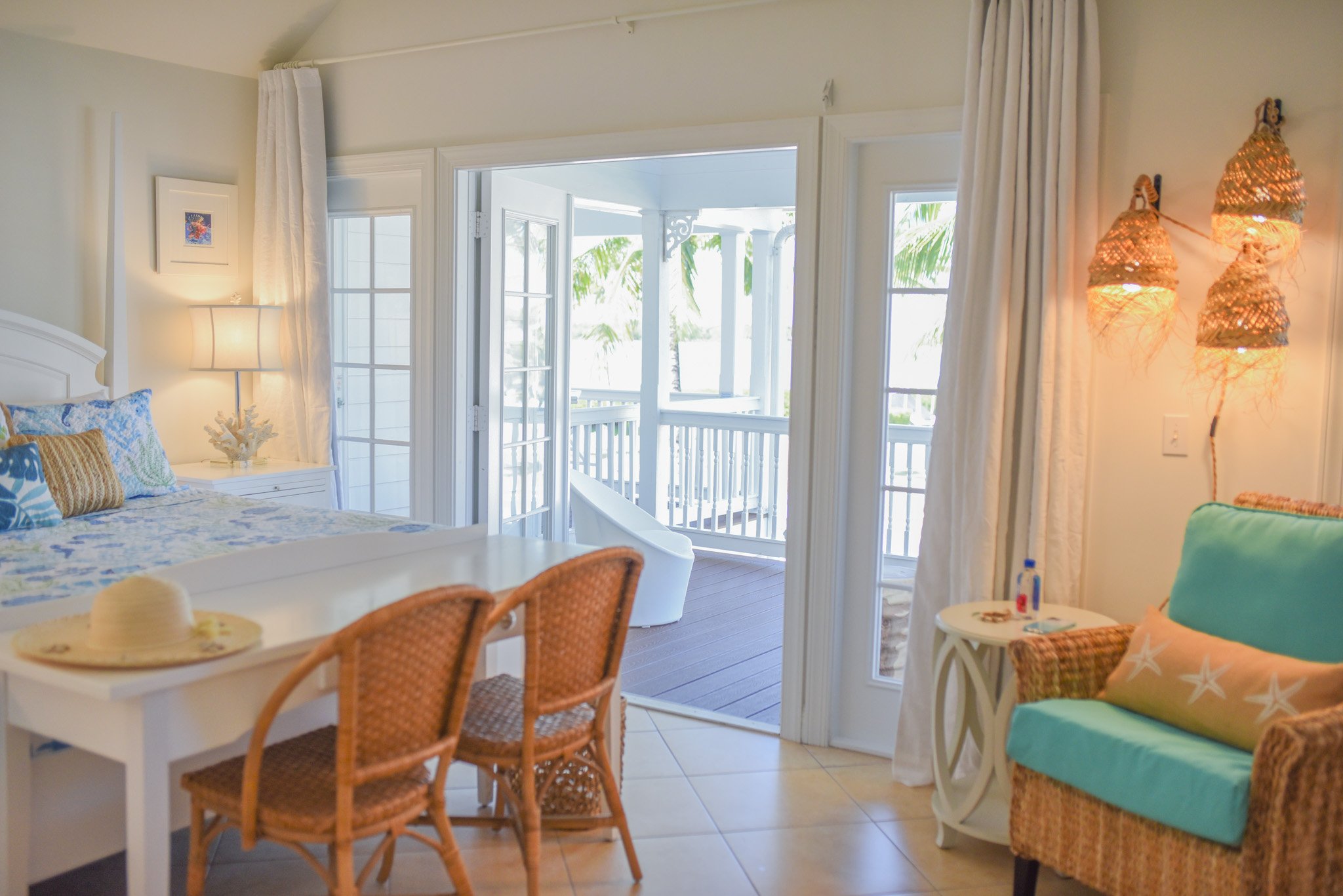 Interior of a beachside hotel room with a bed, a balcony with outdoor furniture, and tropical decor.