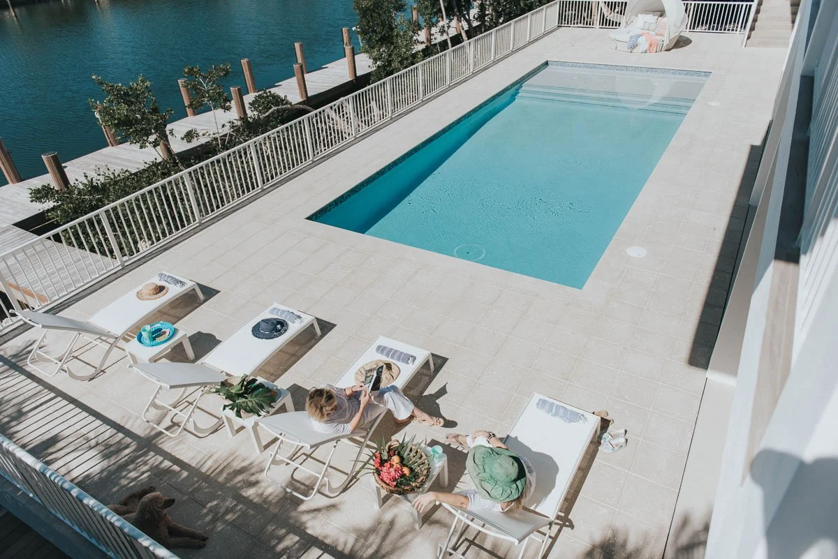 A rooftop pool area with lounge chairs, a woman sitting and reading, a person in a green hat, and a view of water and trees beyond the railing.