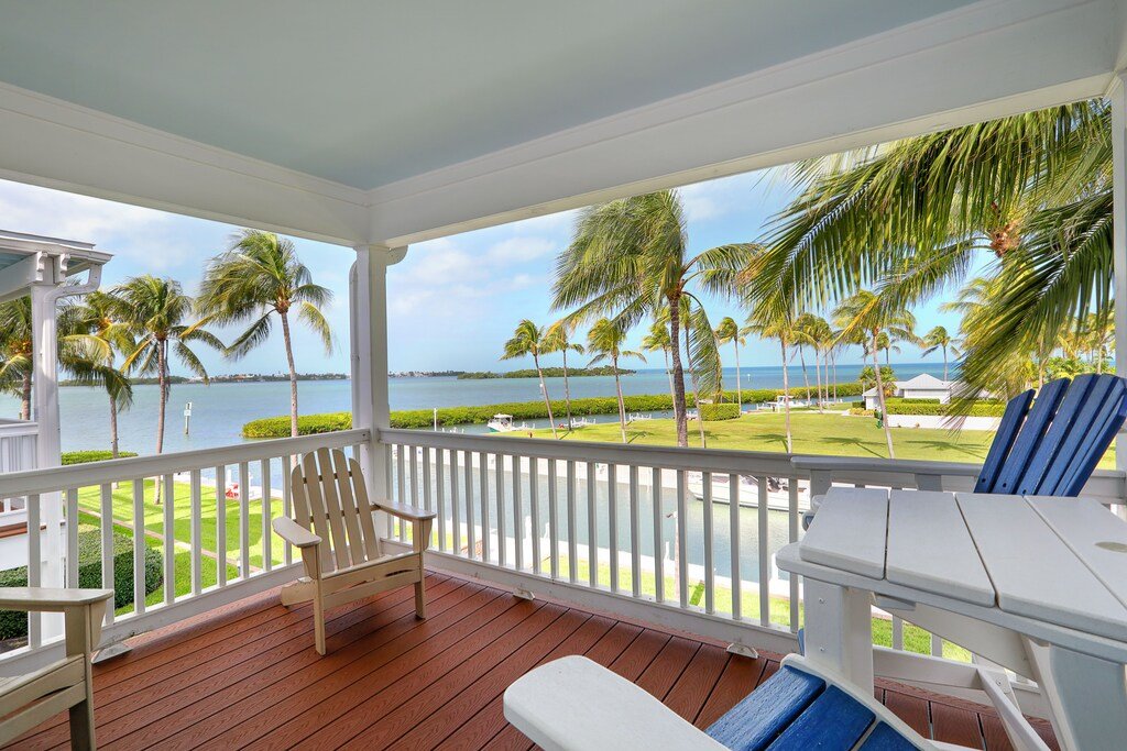 View of a porch overlooking a tropical landscape with palm trees, a swimming pool, water, and a bright blue sky.