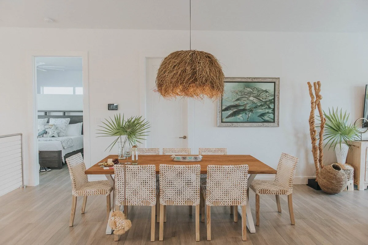 A coastal-themed dining room with a wooden table and eight woven chairs. Two large green palm leaves in vases are on the table. A large, rustic, woven pendant light hangs overhead. In the background, there's a bedroom with a bed decorated with towels
