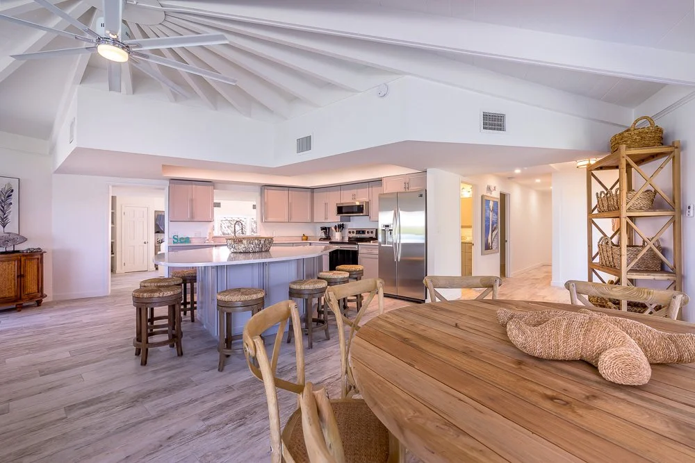 Open-concept kitchen and dining area with a circular wooden table, rattan dog-shaped pillow, and bar stools at a kitchen island with white cabinets and stainless steel appliances.