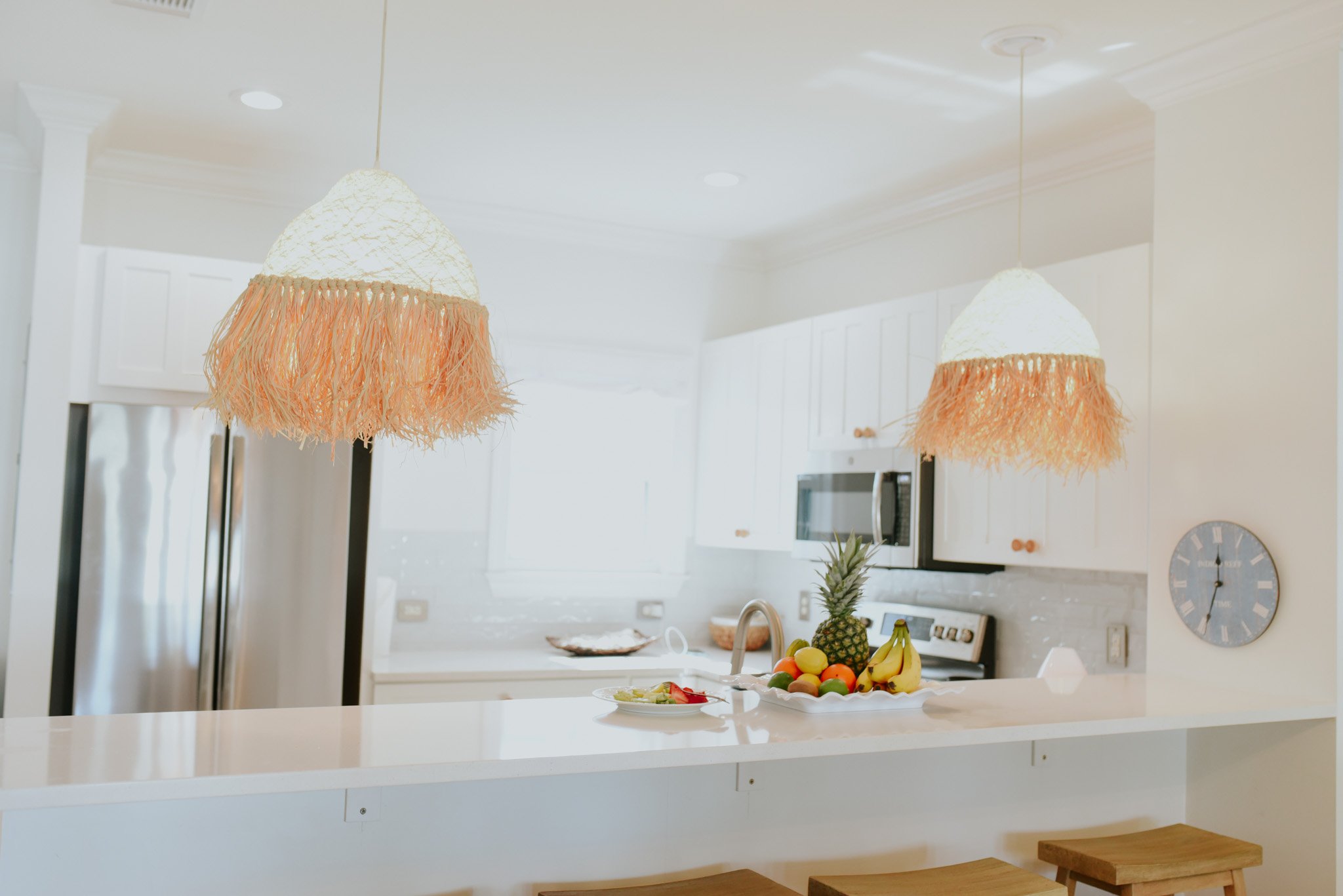 Bright kitchen with two hanging lamps with beige fringed shades, a white countertop with a fruit display including a pineapple, bananas, and other fruit, and a wall clock on the right.