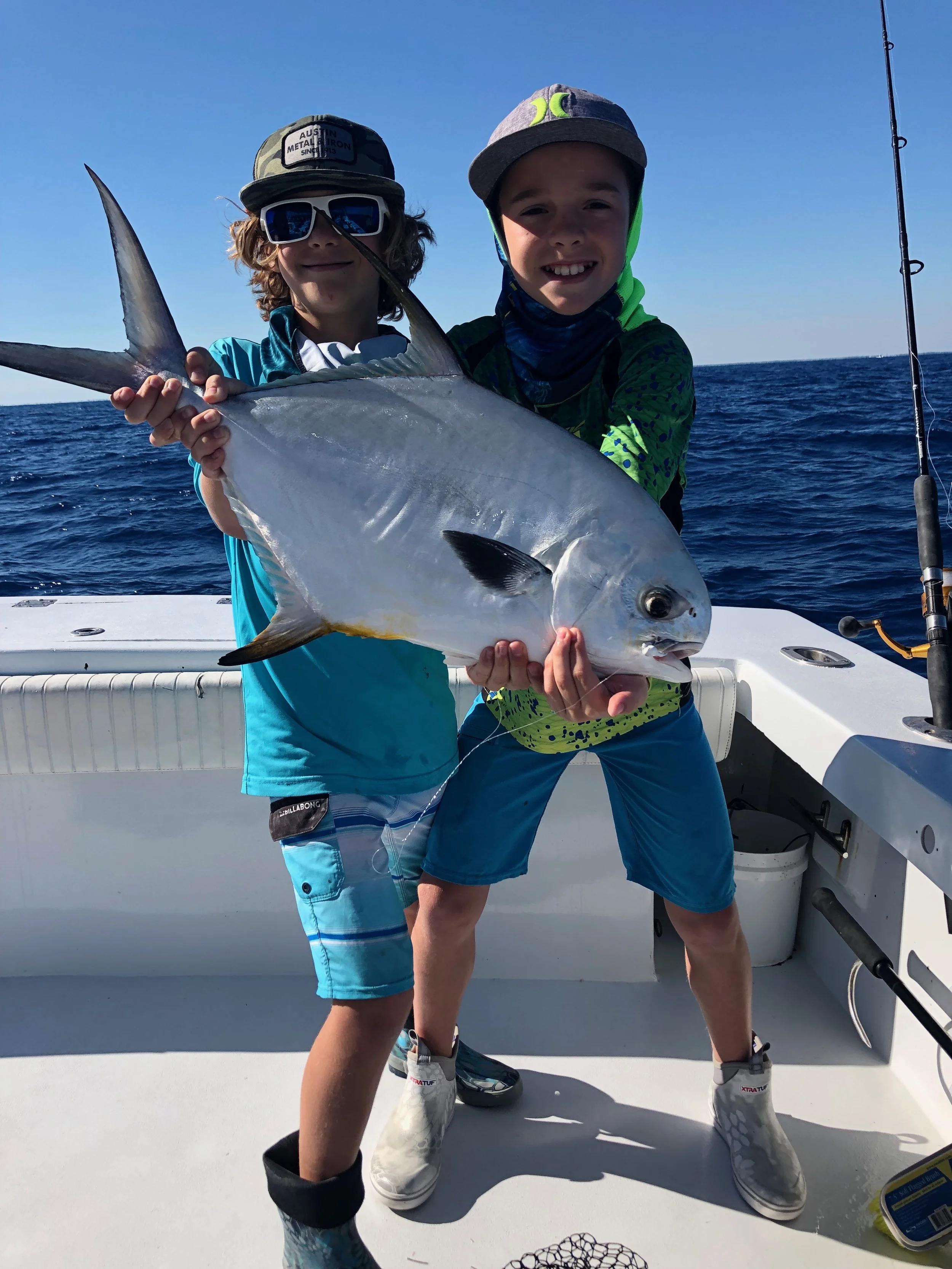Two boys on a boat holding a large fish, with the ocean in the background during a sunny day.