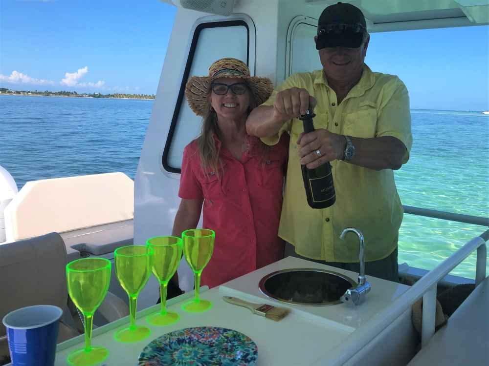 A woman and man on a boat celebrating, with the woman wearing a wide-brimmed hat and the man opening a bottle of champagne, four green champagne glasses on the table, and a view of water and a distant shoreline in the background.