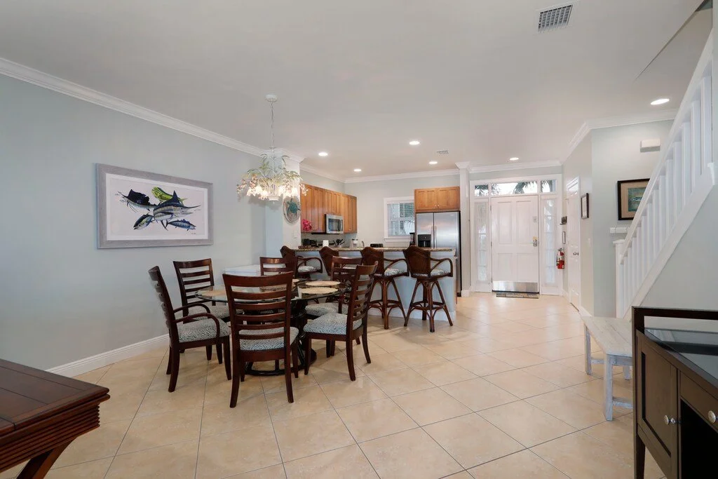 Open concept living space with a dining table, kitchen island with barstools, stainless steel refrigerator, light-colored tile flooring, and light pastel walls.
