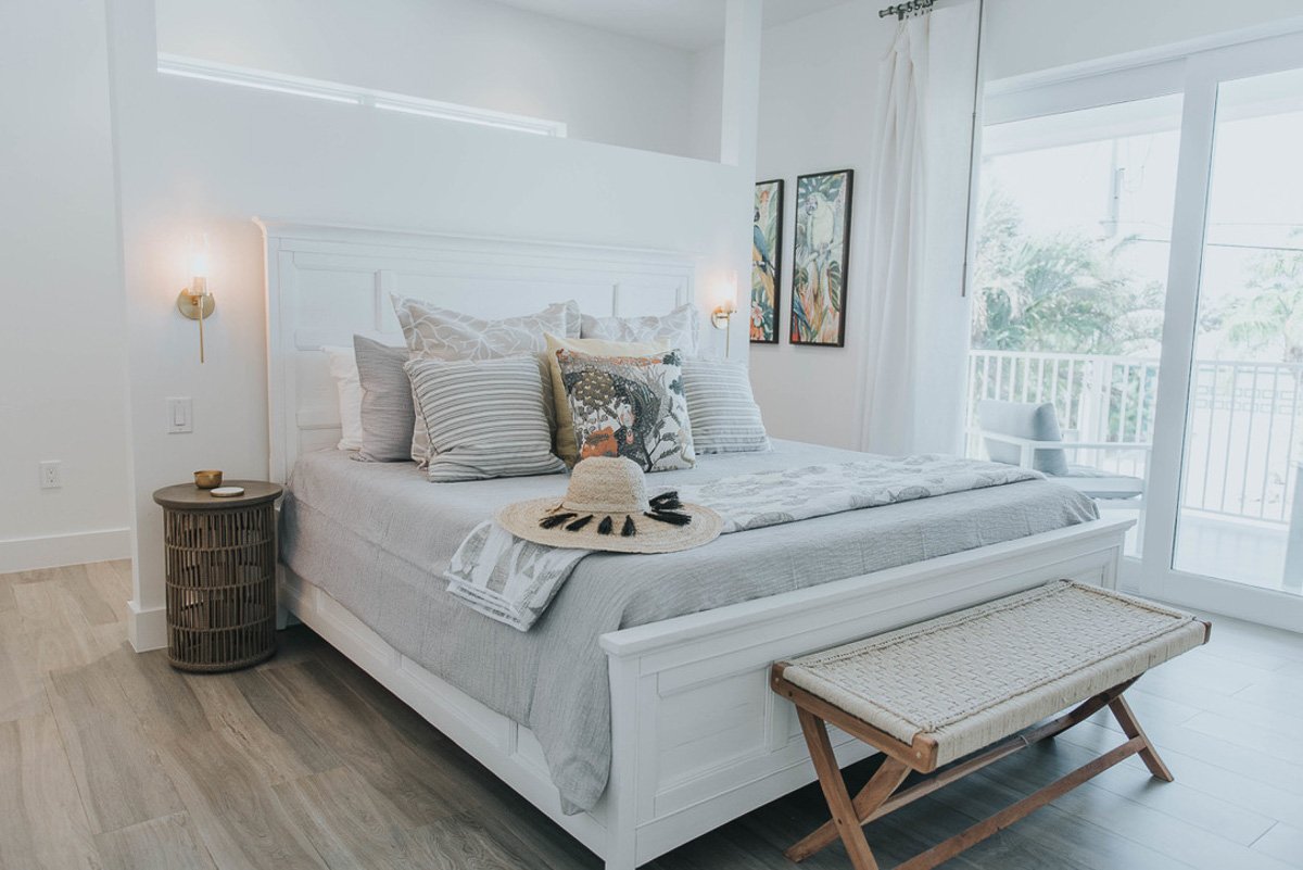 Bright bedroom with a white bed, multiple pillows, and a hat with black tassels on a straw hat, placed on the bed. There's a wooden bench at the foot of the bed, a small round wicker side table, and three framed colorful bird artwork on the wall. Lar