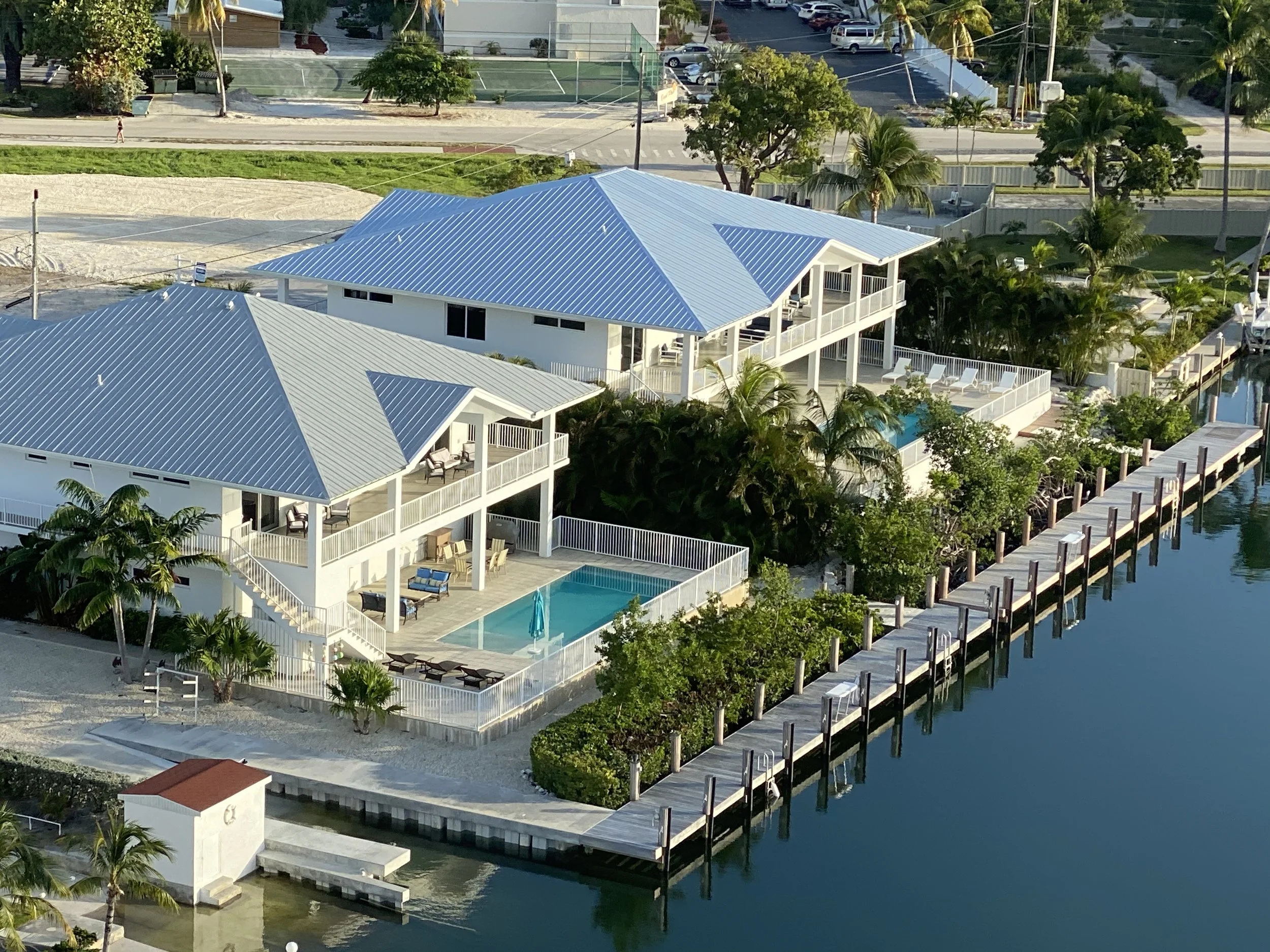 Aerial view of a waterfront property with two white houses, each with a blue metal roof. One house features a swimming pool on the patio with lounge chairs. The property is surrounded by lush greenery and a waterway with a dock. Streets and parking l
