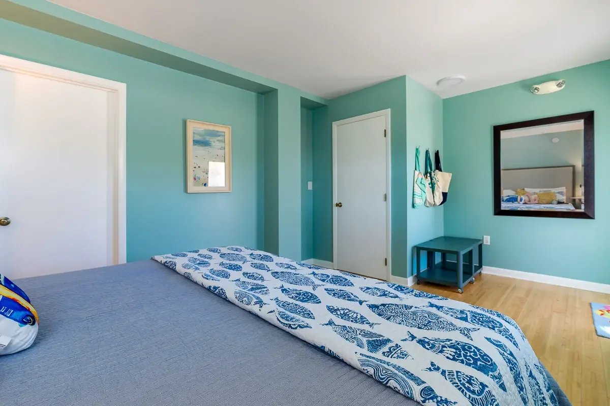 A bedroom with aqua-colored walls, a bed with blue and white patterned bedding, a framed artwork, a mirror reflecting the bed, and a small table with hanging bags.