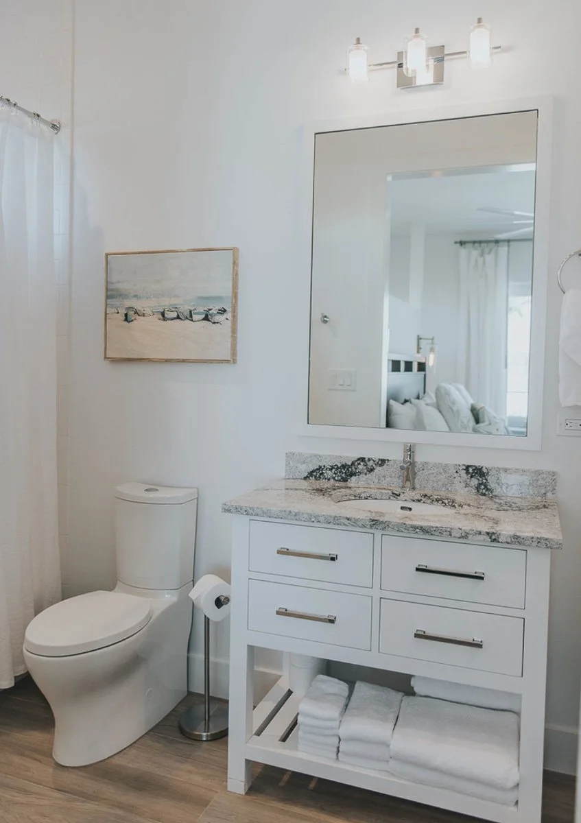 Bathroom with white walls, a toilet, a white vanity with a granite countertop, a large mirror, a light fixture, and a small painting of a beach scene. There's a towel rack with white towels and a entry door reflected in the mirror.