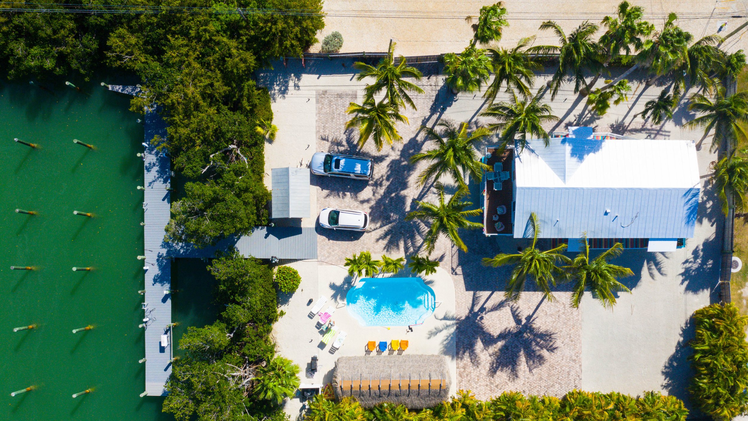 Aerial view of a backyard with a swimming pool, lounge chairs, tall palm trees, a house with a white roof, a parking area with two cars, and a large green sports court surrounded by greenery.