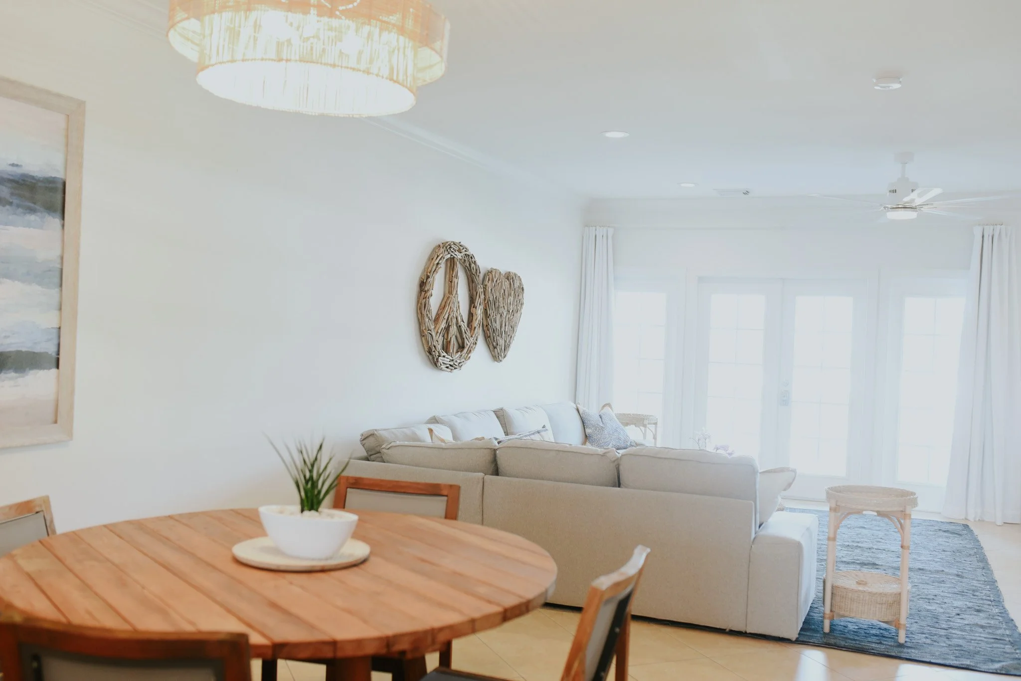 Bright living room with white walls, beige sofa, round wooden dining table with a white plant pot, artwork on the wall, and large windows with white curtains.
