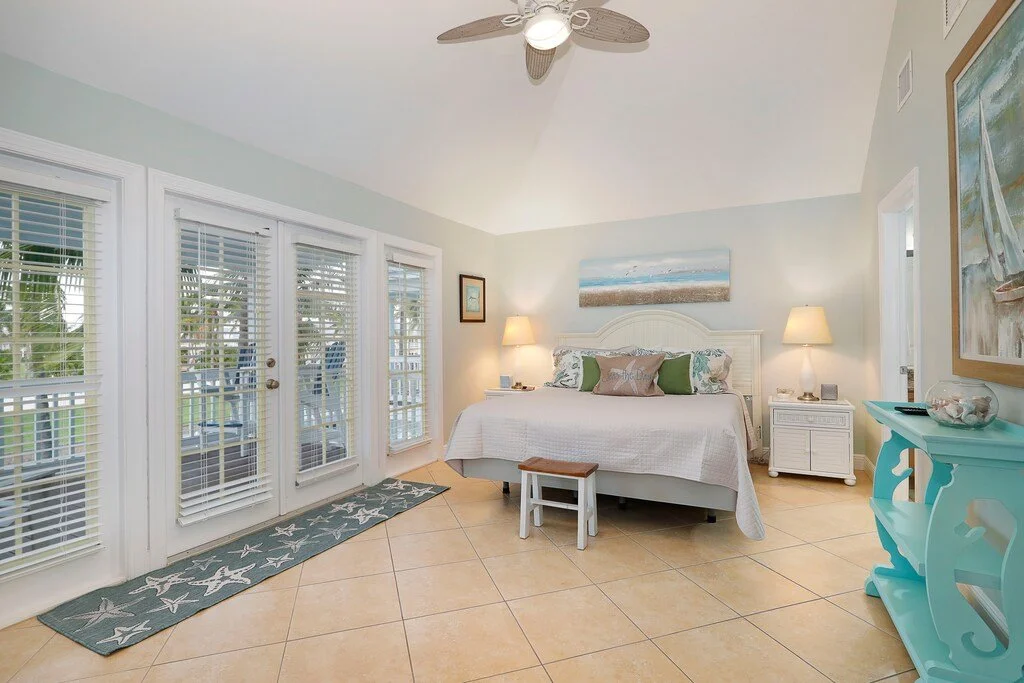 Bright bedroom with a white bed, nightstands with lamps, glass door with blinds leading outside, beach-themed decor, light blue walls, ceiling fan, and tiled floor.