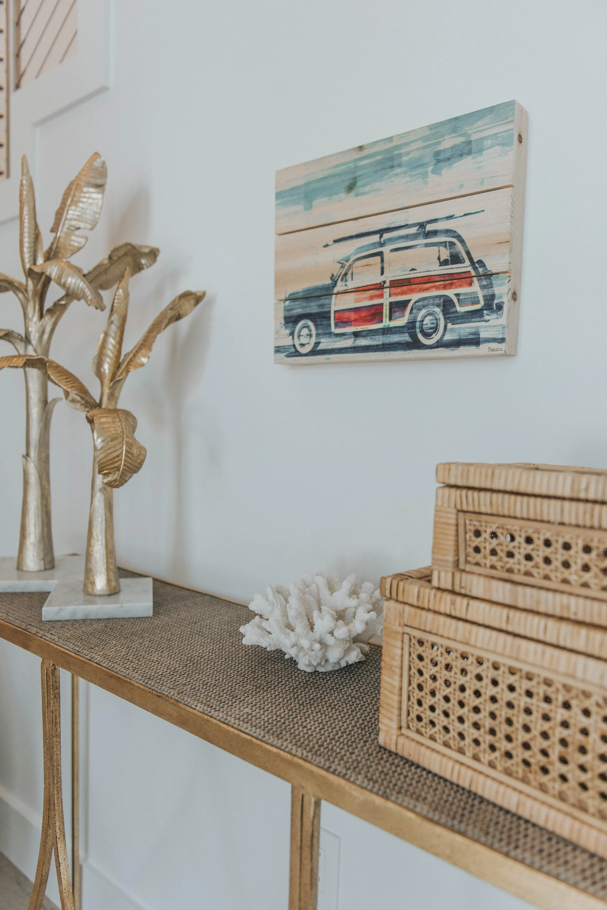 Decorative houseplant, coral piece, and rattan boxes on a golden console table, with a wooden wall art of a vintage car hanging above.