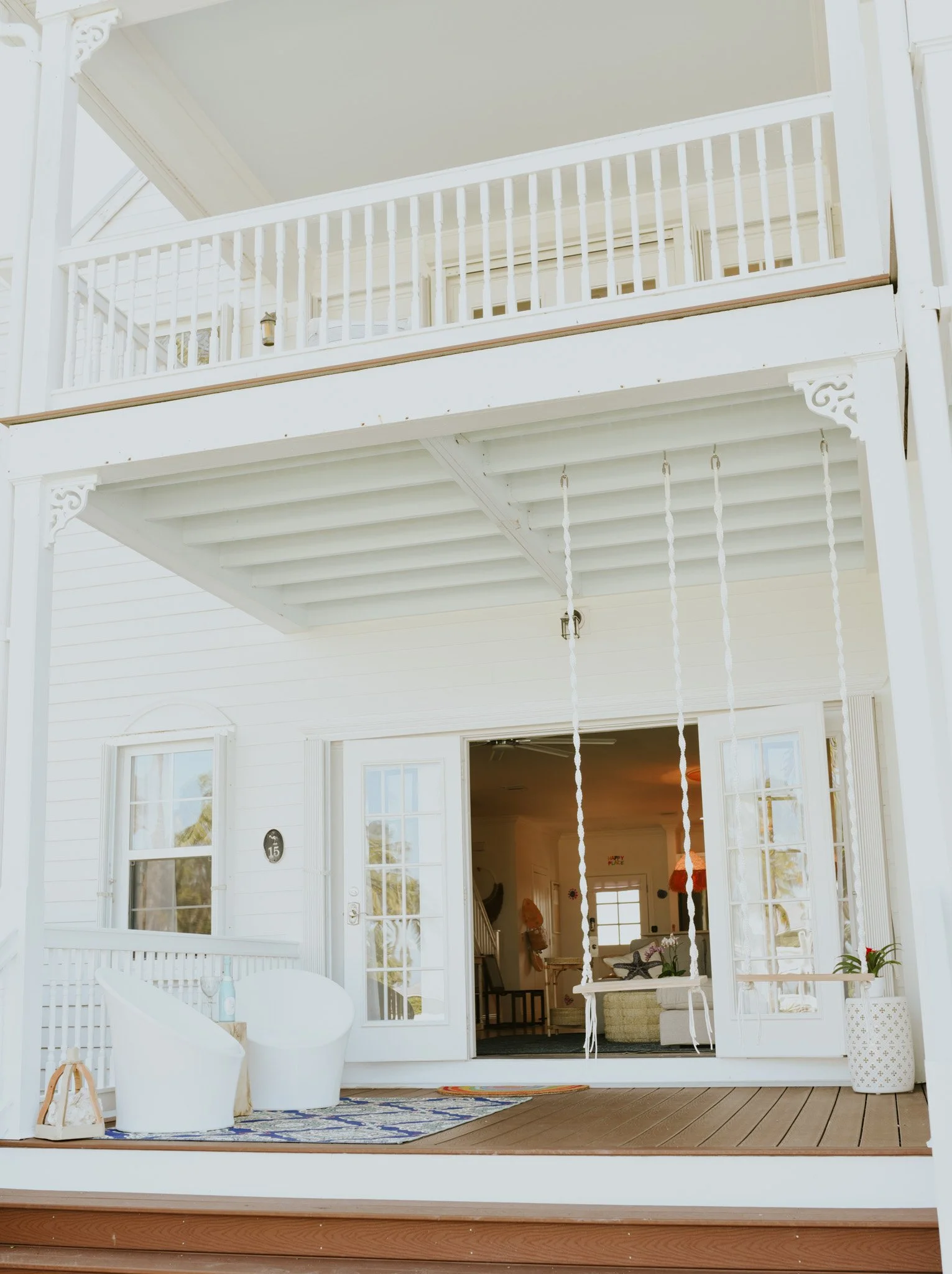 A white two-story house with a front porch featuring hanging swings, glass-paneled double doors, and large windows. Outside, there is a patterned rug, two white chairs, and a potted plant.