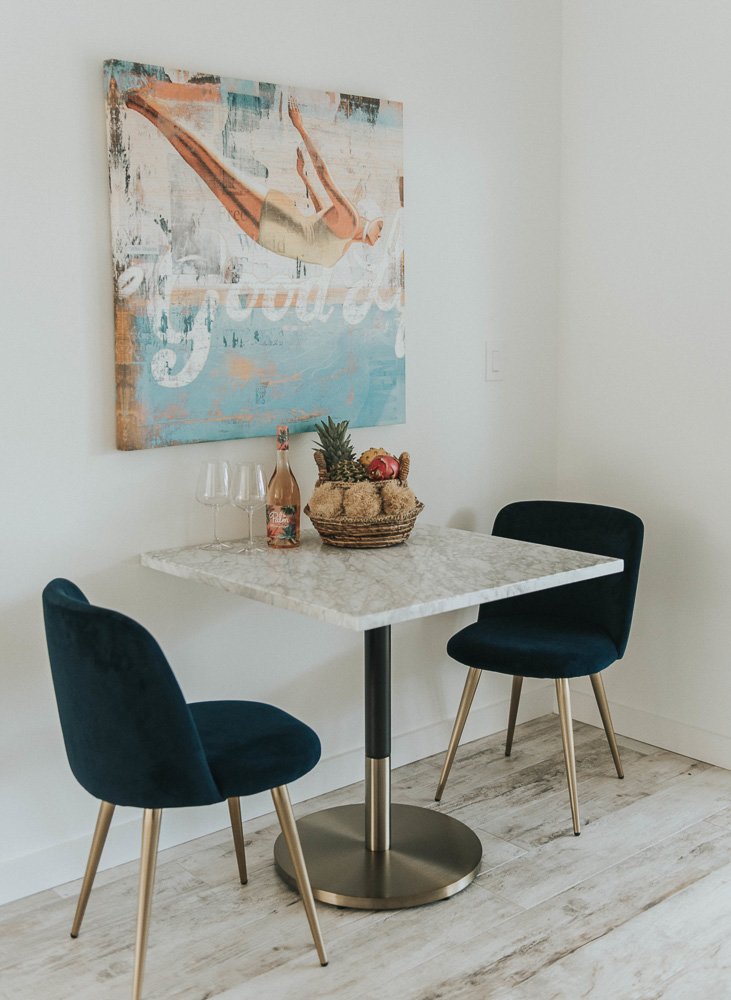 A small dining area with a square marble table, two navy blue velvet chairs with gold legs, a framed artwork of a woman in vintage swimwear hanging on the white wall, a basket of assorted fruits including pineapples and apples, and two wine glasses a